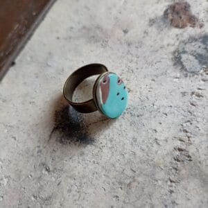 bague ovale couleur turquoise et marron avec un anneau en laiton, posée sur un fond gris.
