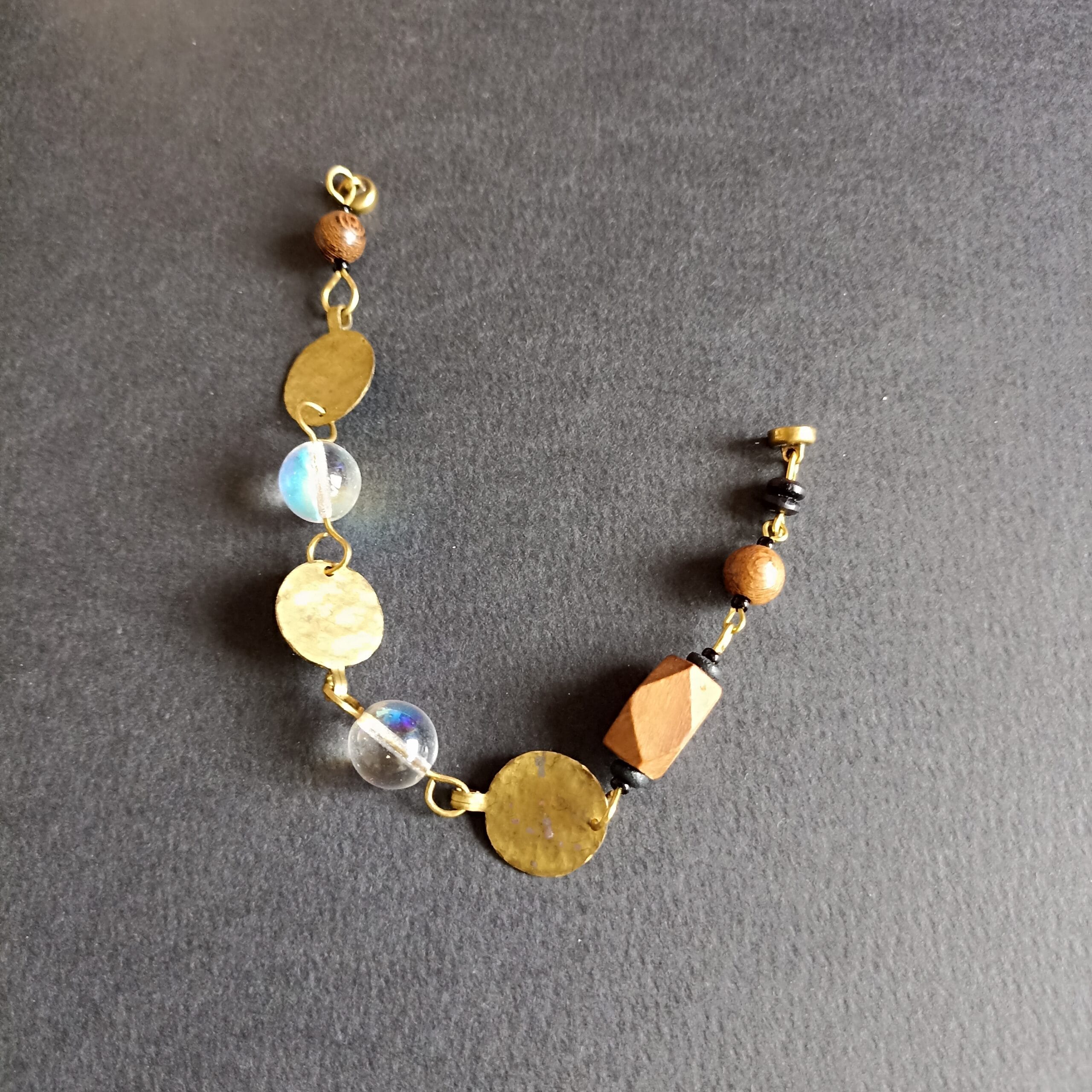 Bracelet Goutte avec perles de bois, verre irisé et laiton martelé posé sur un fond noir.